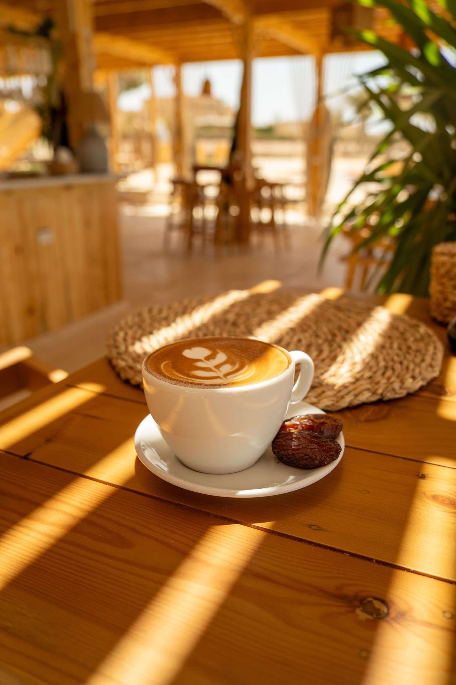 A cappuccino with latte art and a date, on a wooden table striped with sunlight