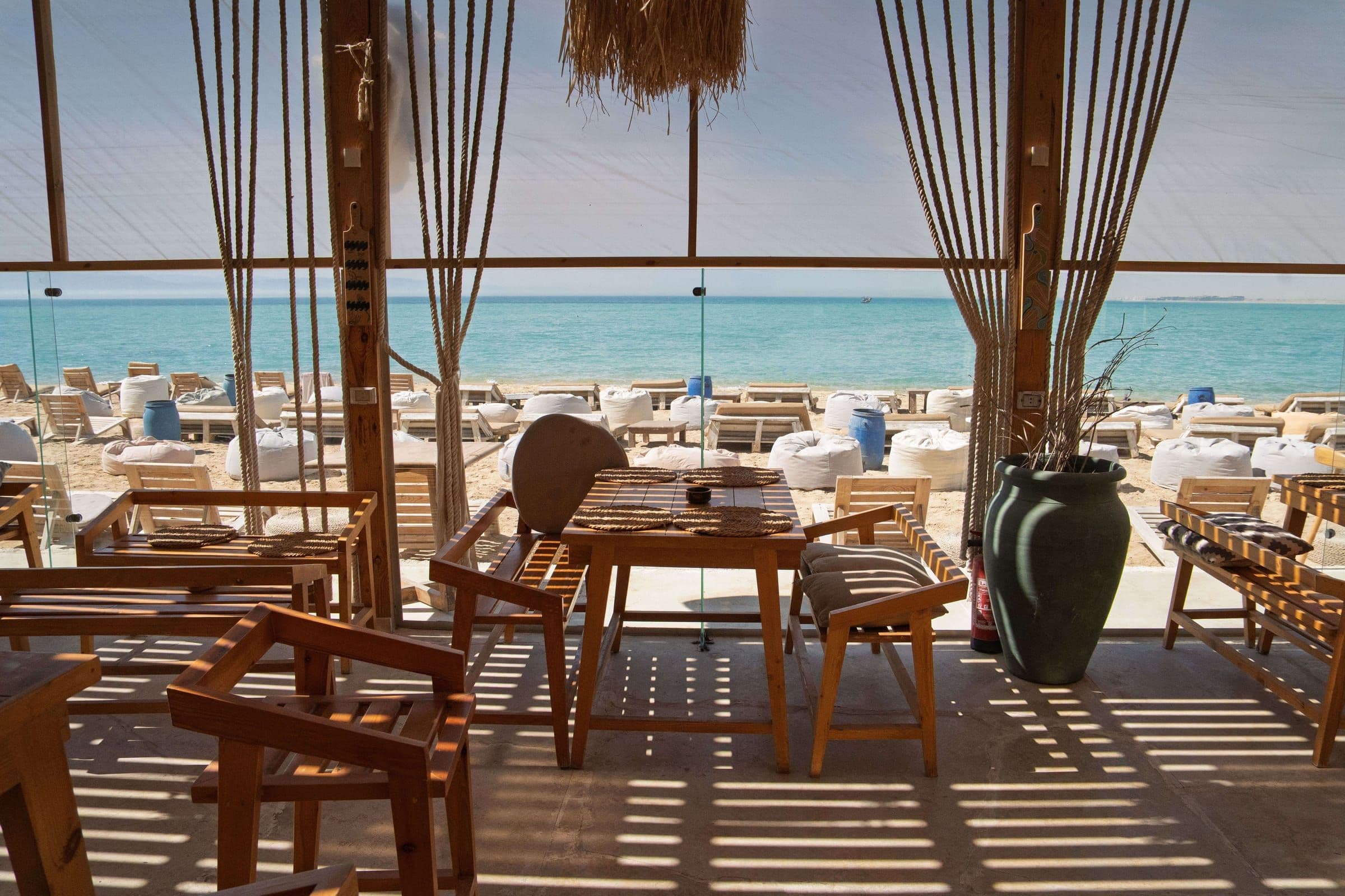 Wooden tables and chairs under the shade of the beach club, looking out to turquoise water with loungers on the sand