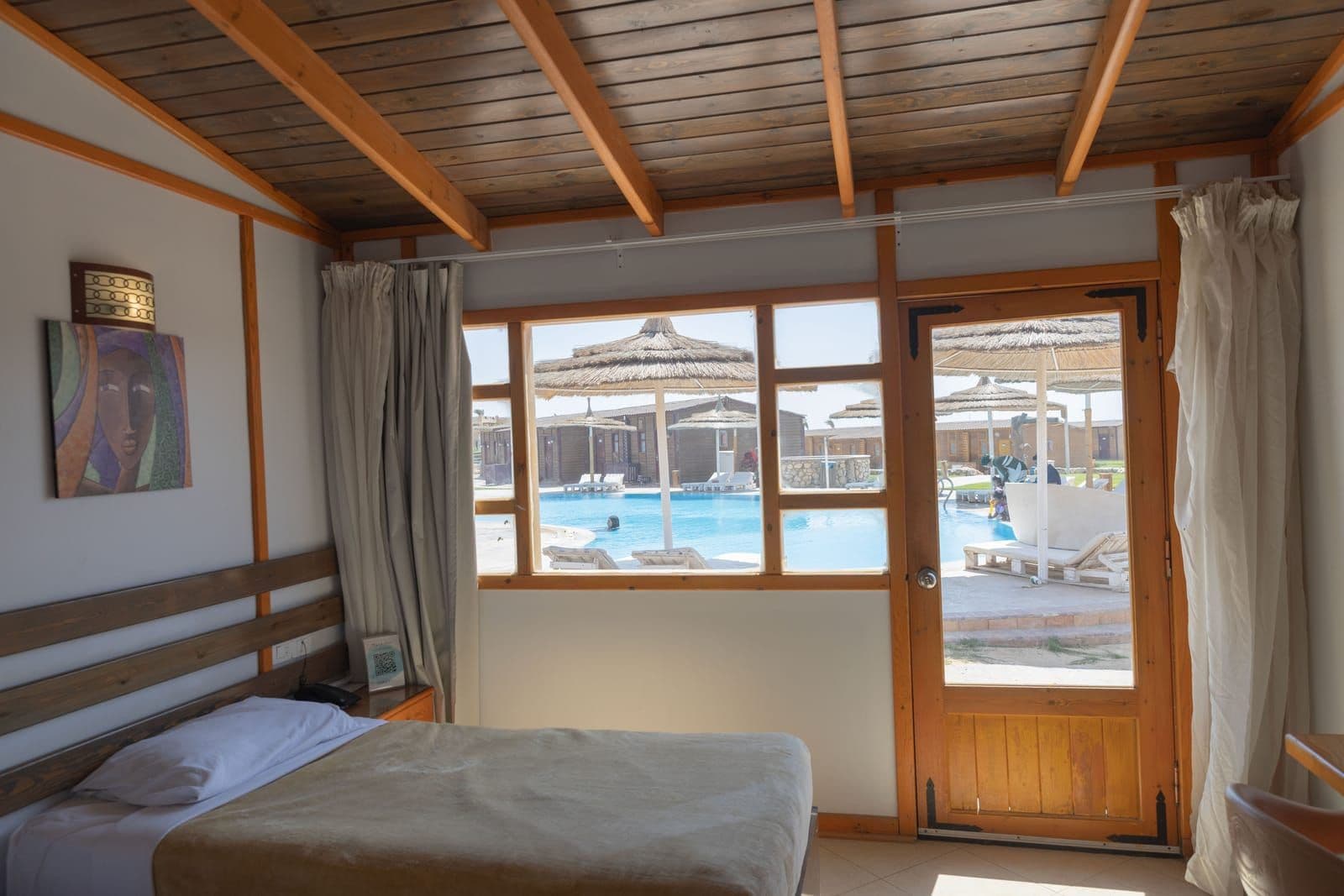 Bungalow double bed beneath a timber-beamed ceiling, the pool and its thatched parasols visible through the window and the glazed door