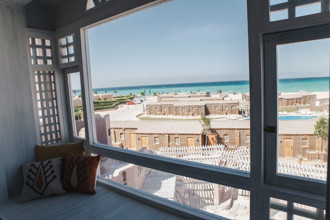 The turquoise sea and the beach seen over the resort rooftops from a cushioned window seat inside a Special Deluxe room