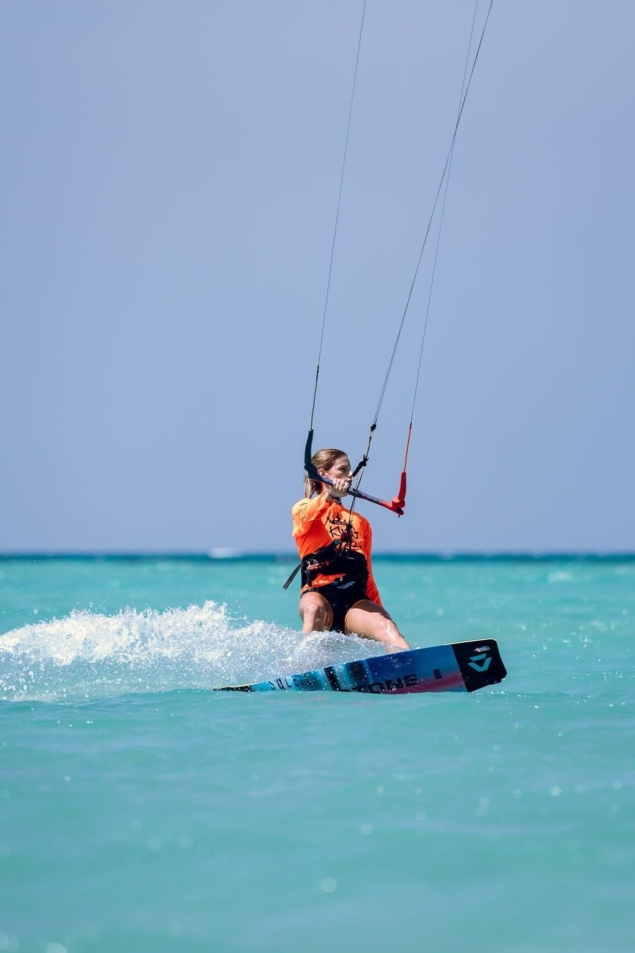 A kitesurfer in an orange event jersey riding towards the camera through shallow turquoise water