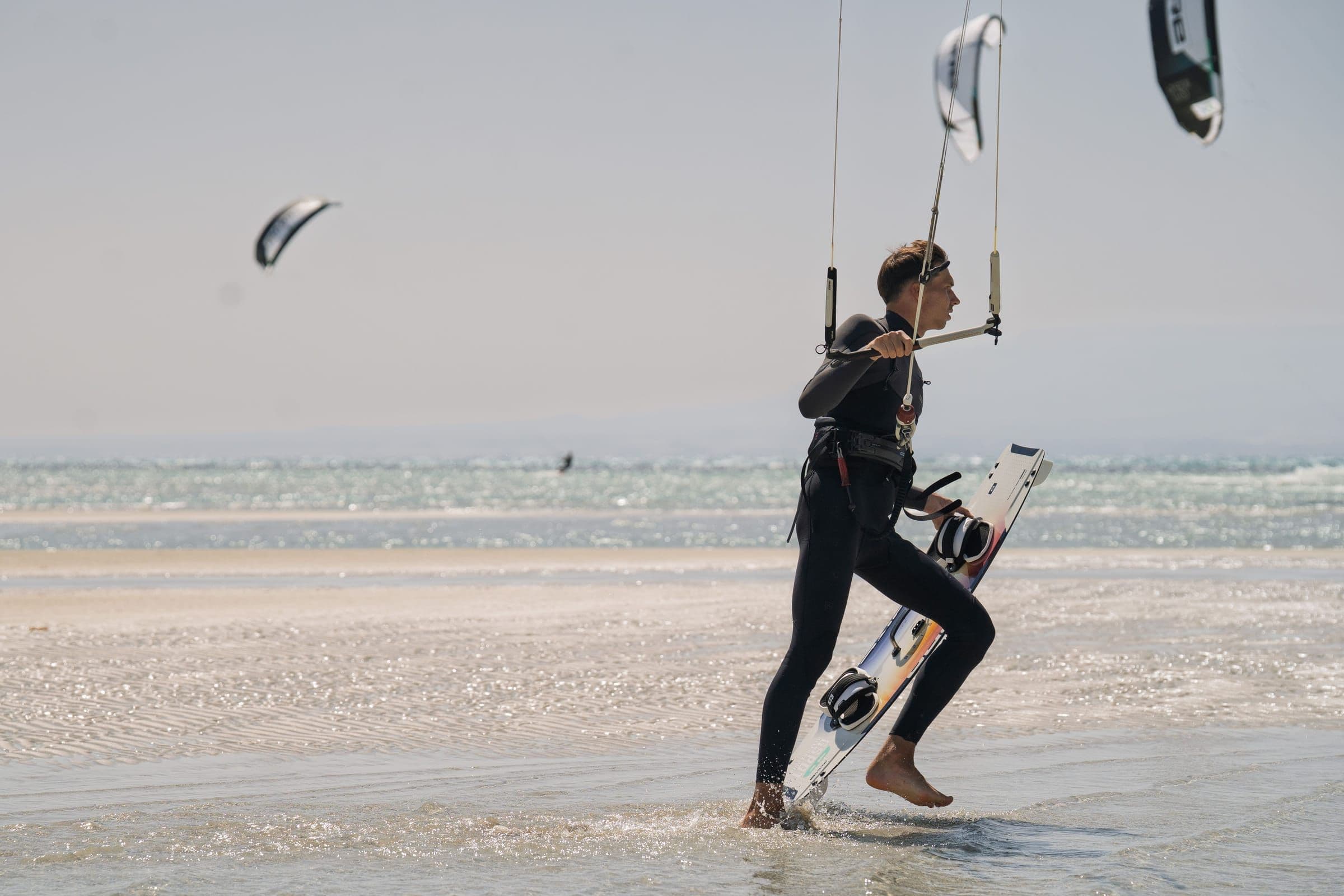 A rider carries a kiteboard into the shallow water while holding the control bar.