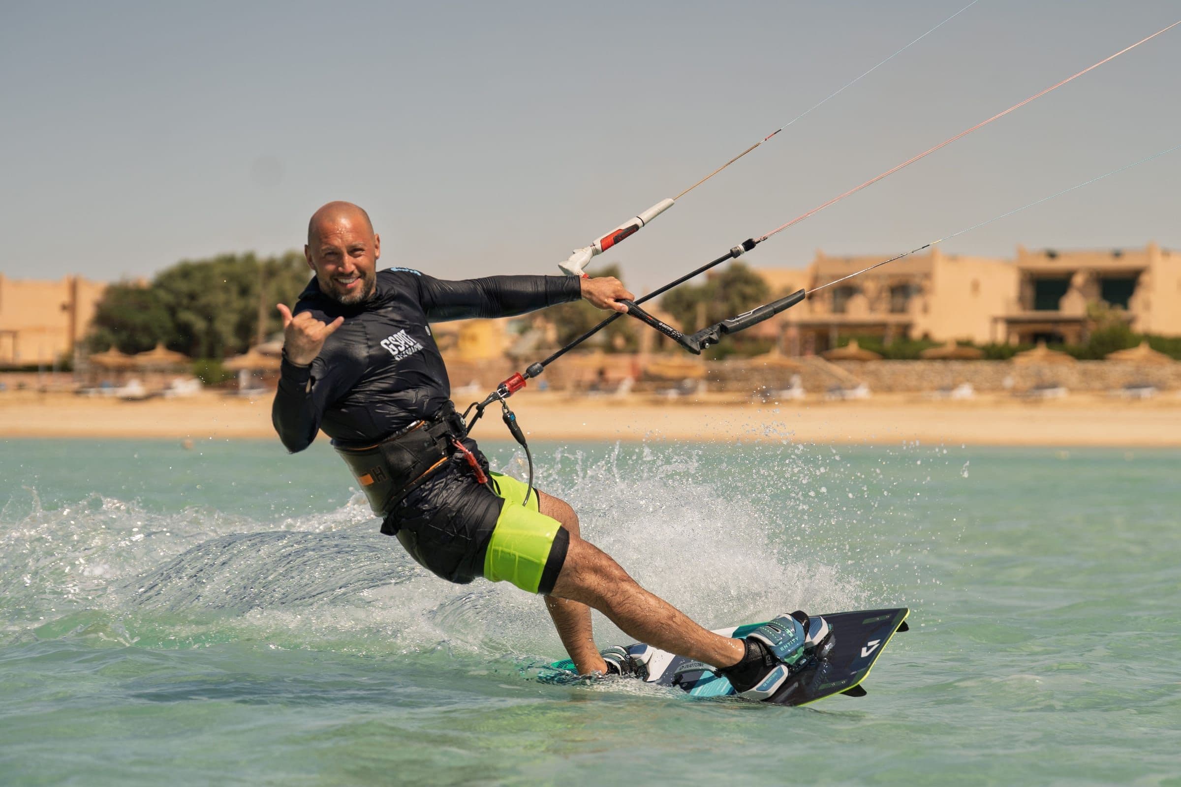 A kitesurfer smiles and makes a shaka sign while riding past the beach.