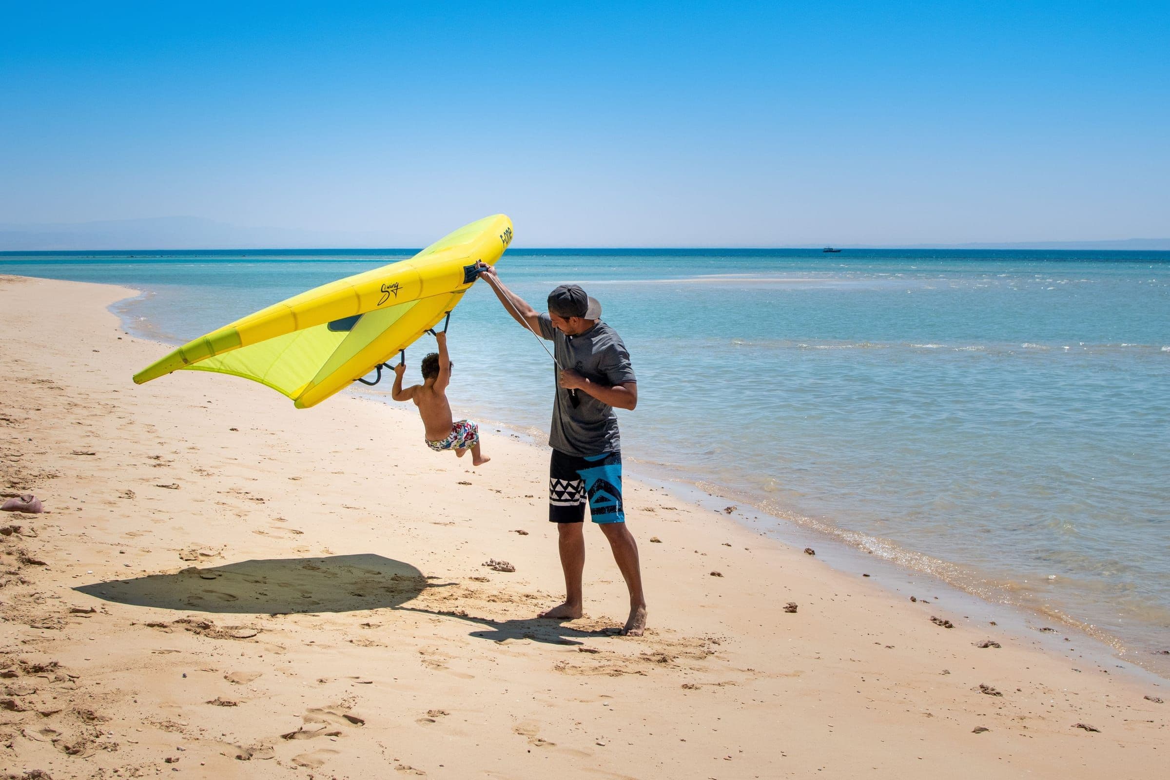 An adult holds a yellow wing while a child swings from its handle on the beach.