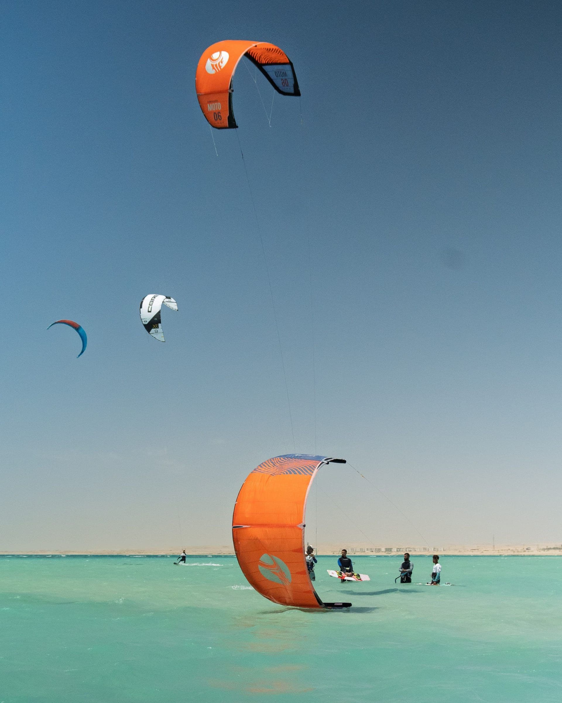 Several kites fill the sky above riders standing in shallow turquoise water.