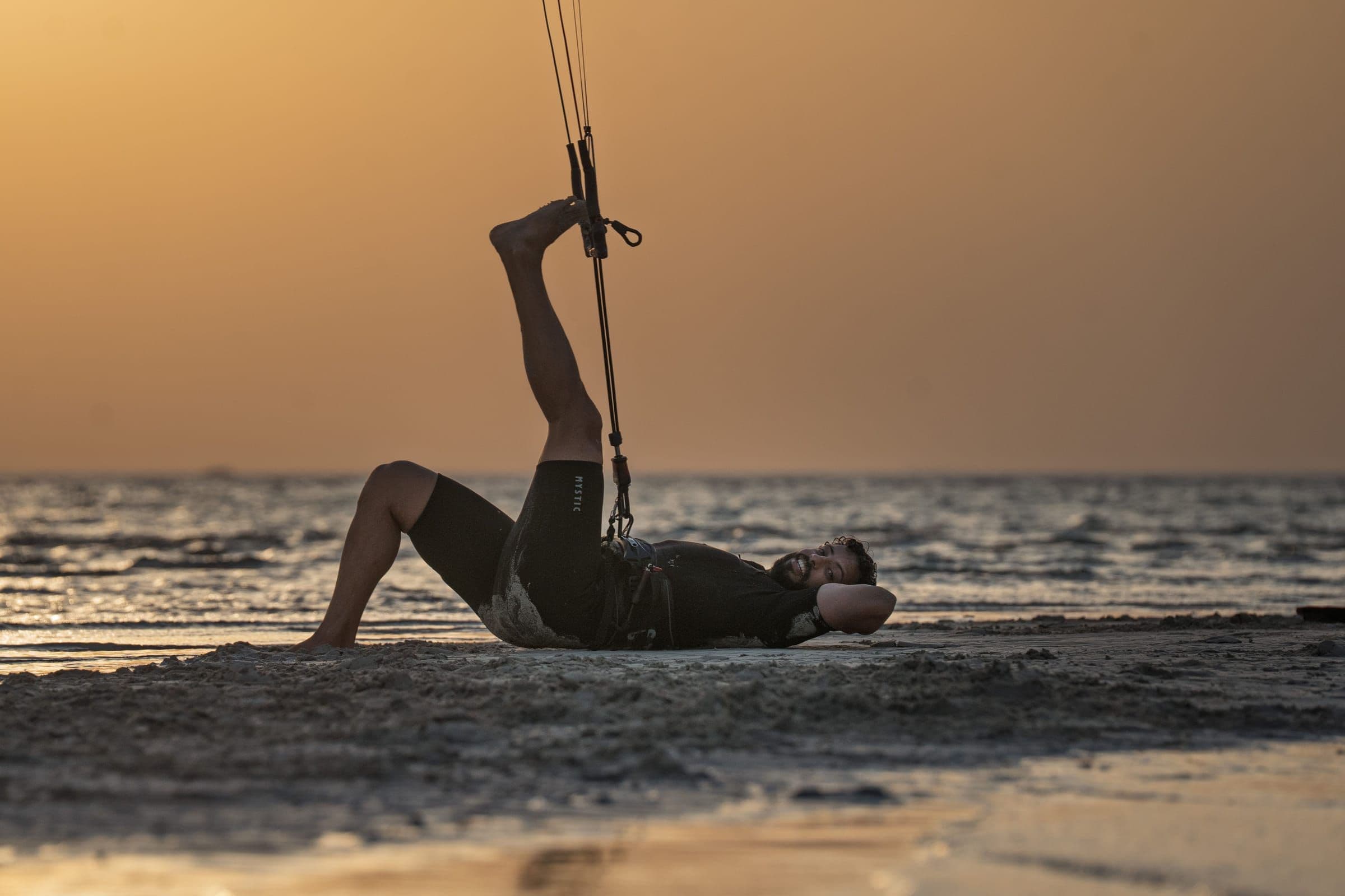 A kitesurfer relaxes on the sand at sunset with one foot resting on the kite bar.