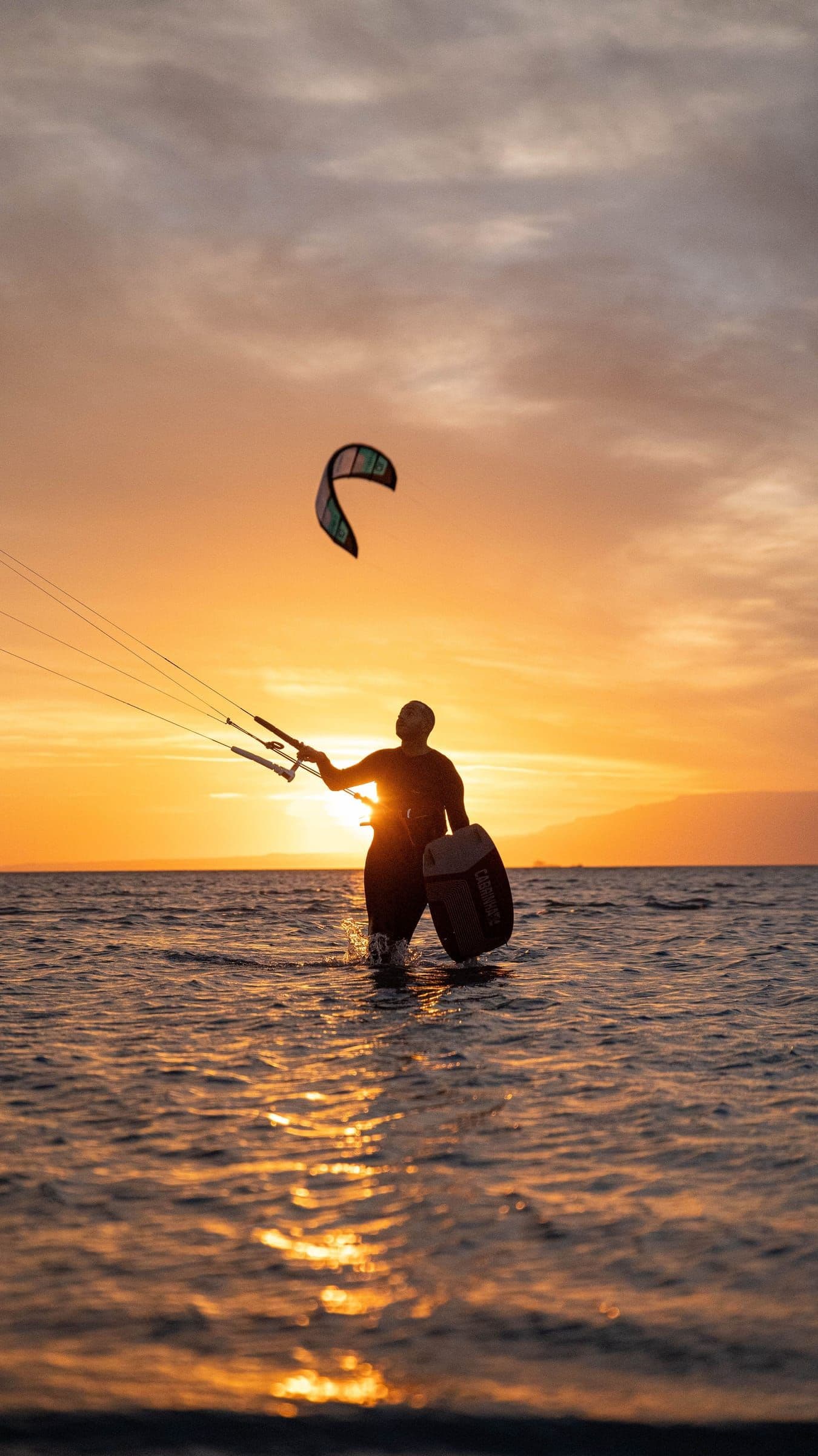 A kitesurfer walks through the water with a board during an orange sunset.